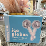 A light blue box of esw beauty ice globes facial tools, featuring an illustration of two pink glitter-coated globes, displayed on a woven table with decorative dried grass and a disco ball in the background.