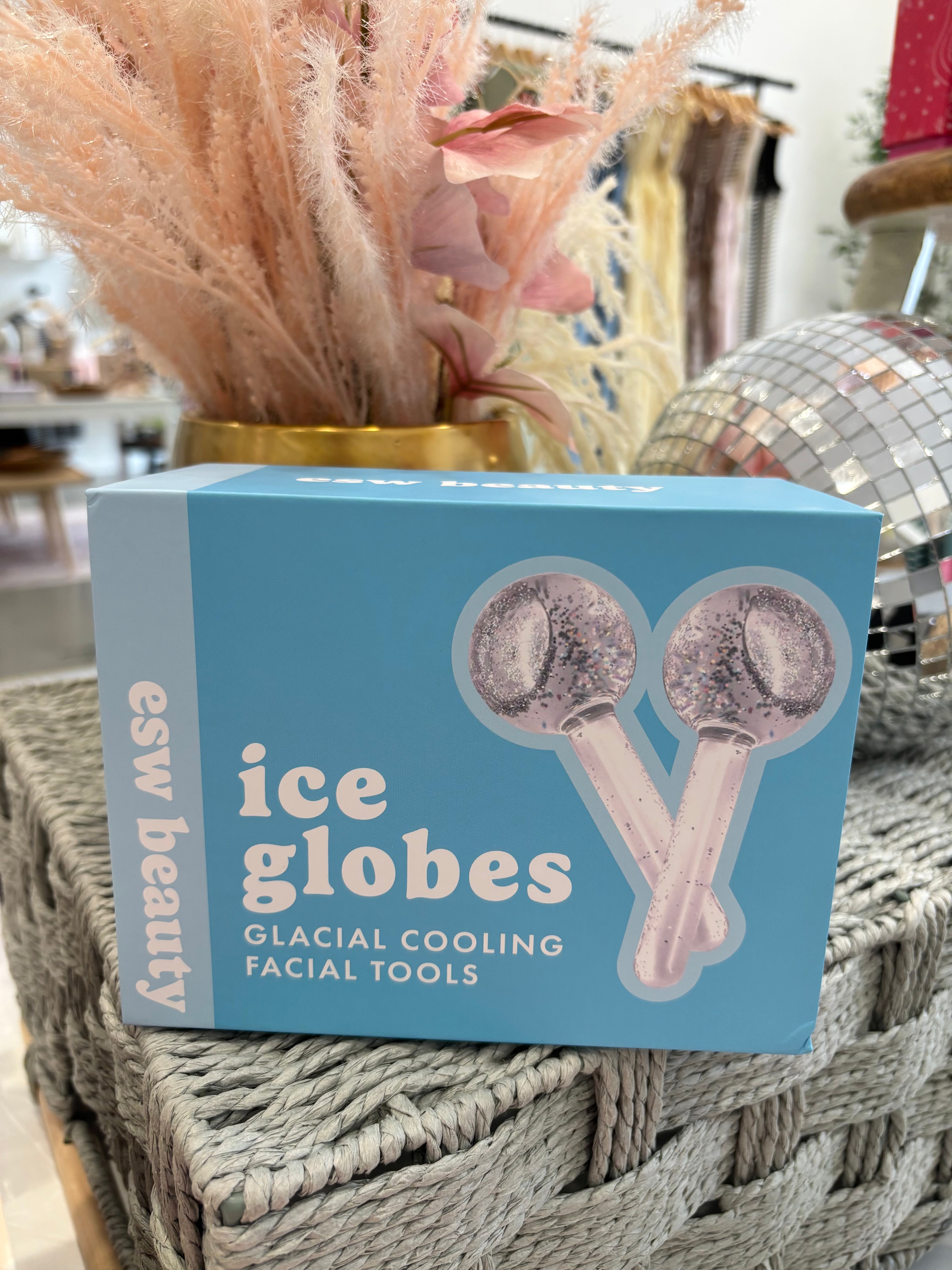 A light blue box of esw beauty ice globes facial tools, featuring an illustration of two pink glitter-coated globes, displayed on a woven table with decorative dried grass and a disco ball in the background.
