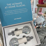 A boxed facial gua sha set from esw beauty, featuring two clear glass massage tools filled with iridescent glitter and white textured handles, presented in an open light blue and white box.