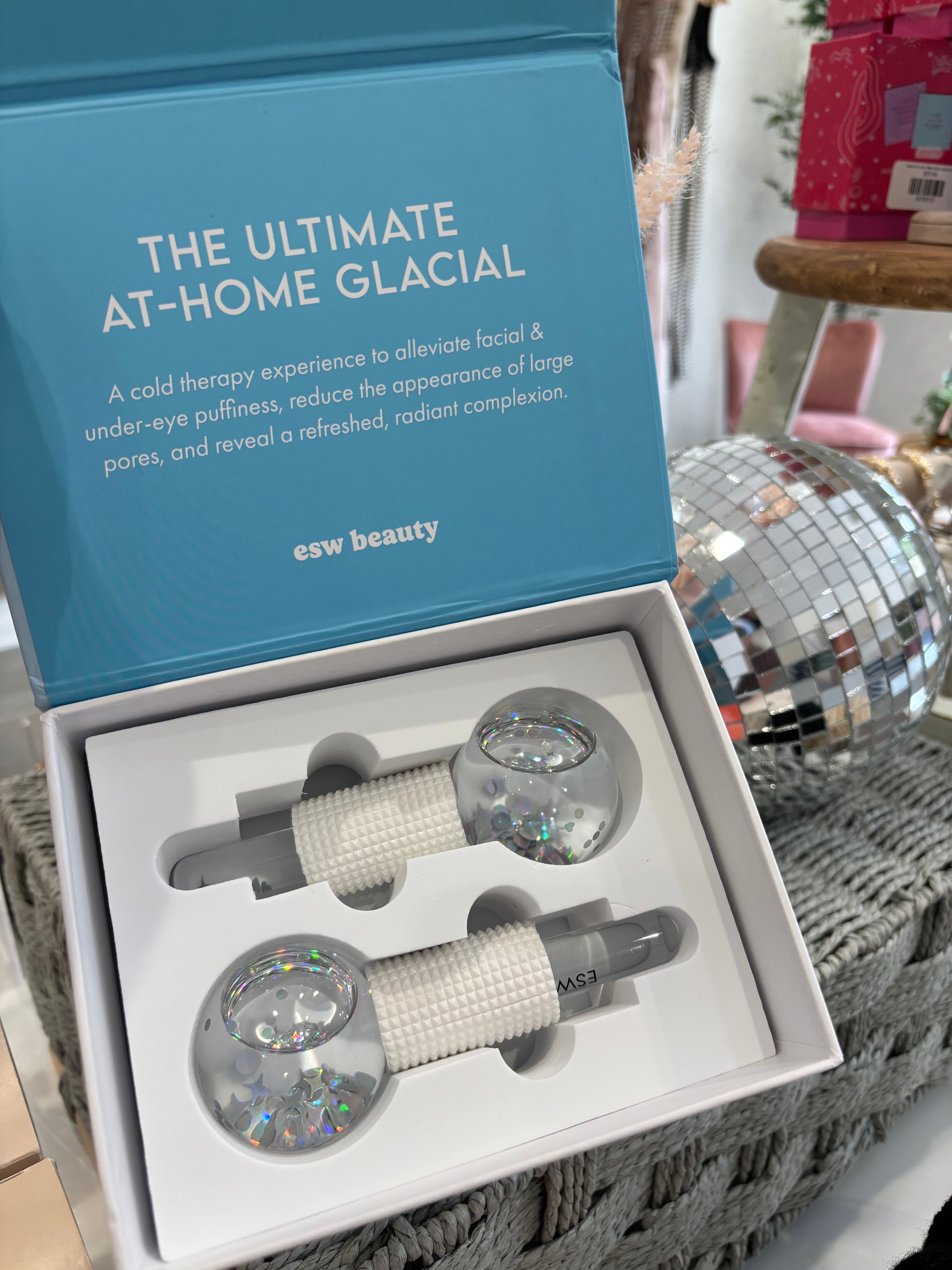 A boxed facial gua sha set from esw beauty, featuring two clear glass massage tools filled with iridescent glitter and white textured handles, presented in an open light blue and white box.