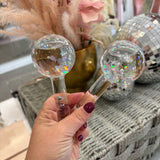 A hand with dark burgundy nail polish holds two clear glass facial massage tools with holographic glitter inside, featuring wooden handles and gold-toned metal accents. The background shows a cozy, styled setting with a woven basket, a gold vase of dried pampas grass, and a disco ball.