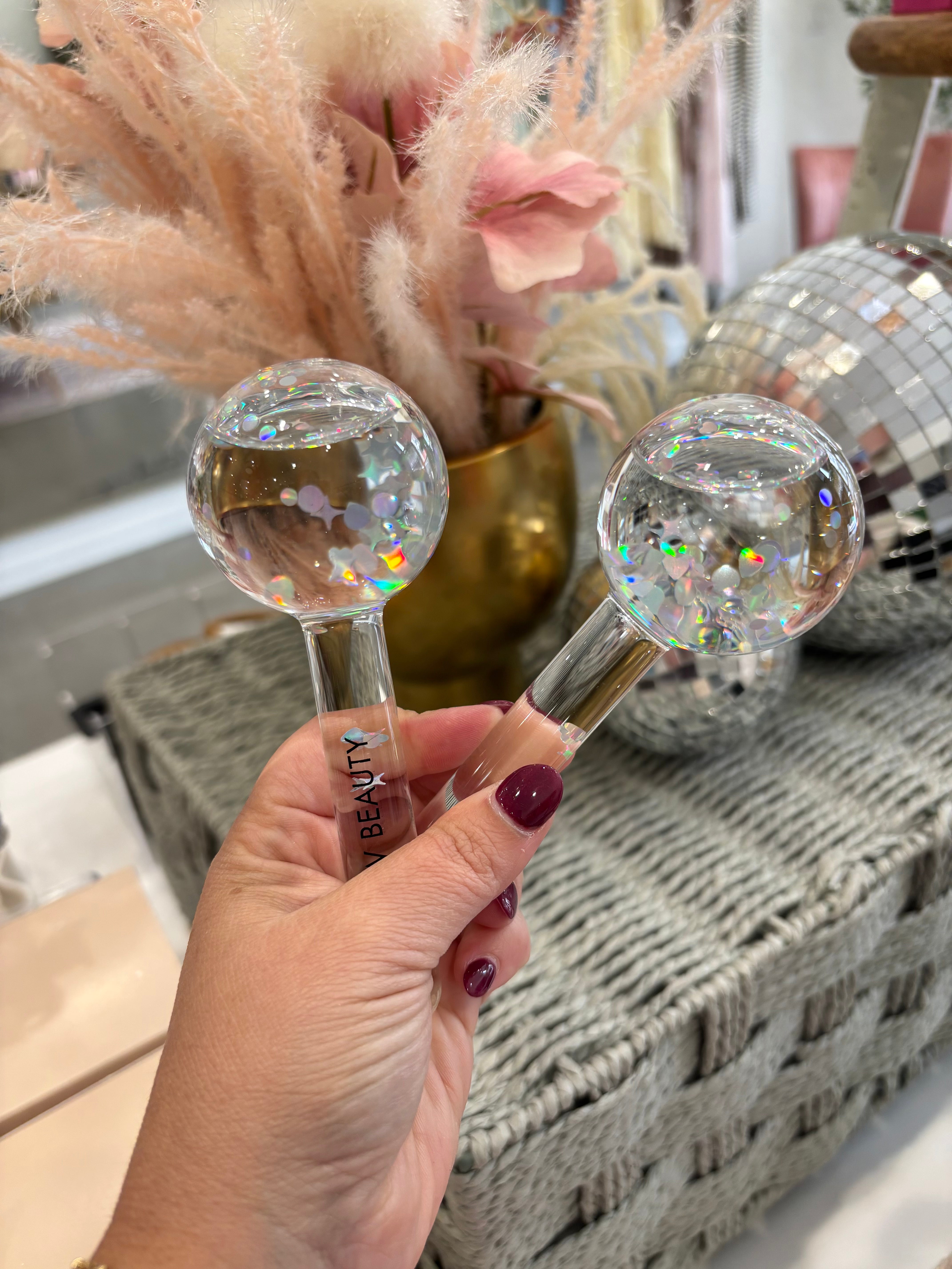 A hand with dark burgundy nail polish holds two clear glass facial massage tools with holographic glitter inside, featuring wooden handles and gold-toned metal accents. The background shows a cozy, styled setting with a woven basket, a gold vase of dried pampas grass, and a disco ball.