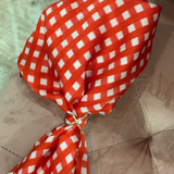 A red and white gingham bow tie with a small gold rhinestone knot, displayed on a brown tufted velvet surface.