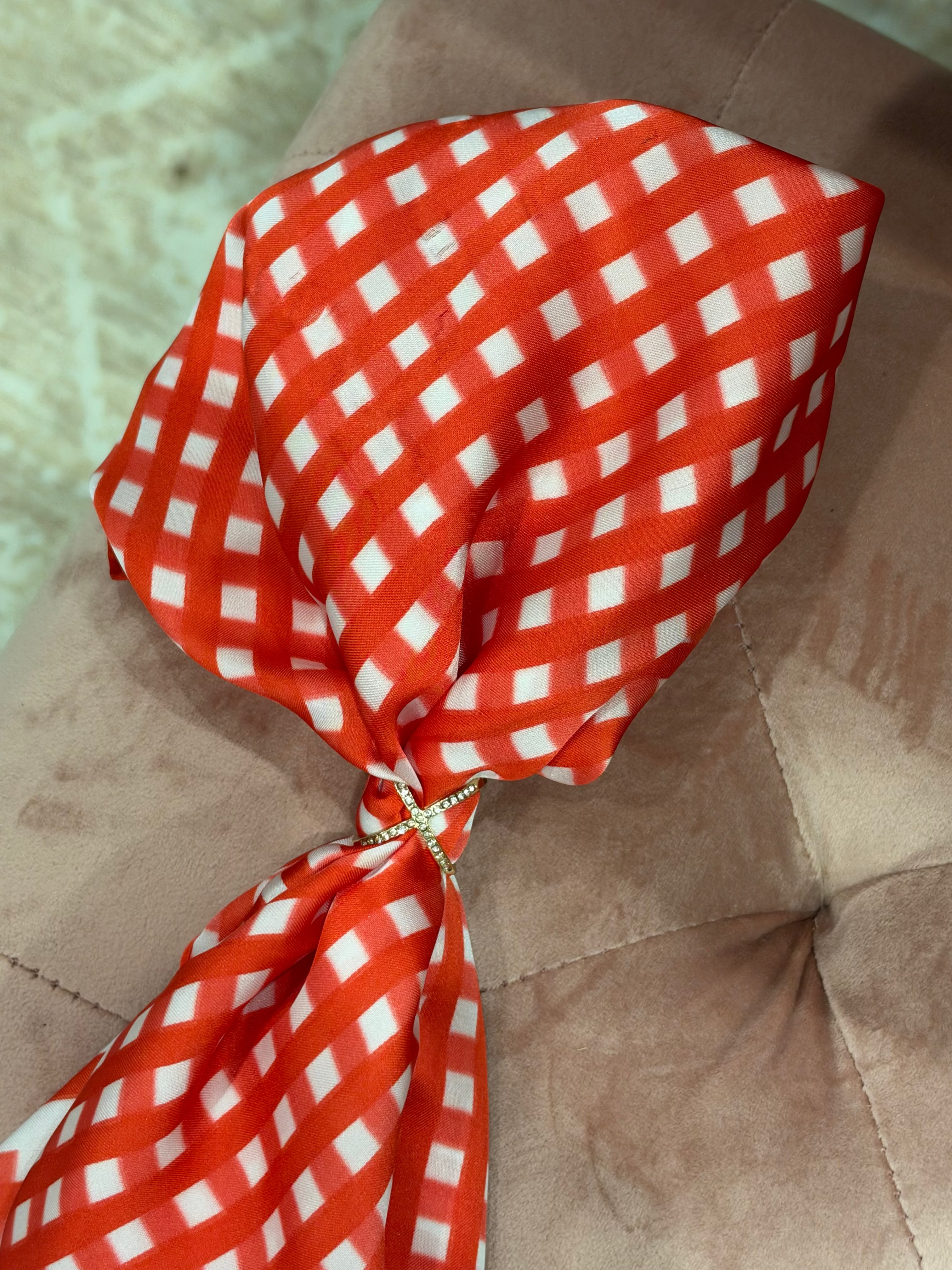 A red and white gingham bow tie with a small gold rhinestone knot, displayed on a brown tufted velvet surface.