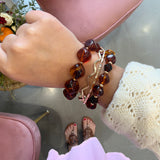 A close-up of a person's wrist wearing multiple bracelets, including amber-colored stone beads and a rose gold chain, with a cream knit sweater sleeve visible. The person is sitting in a pink chair in an indoor setting.