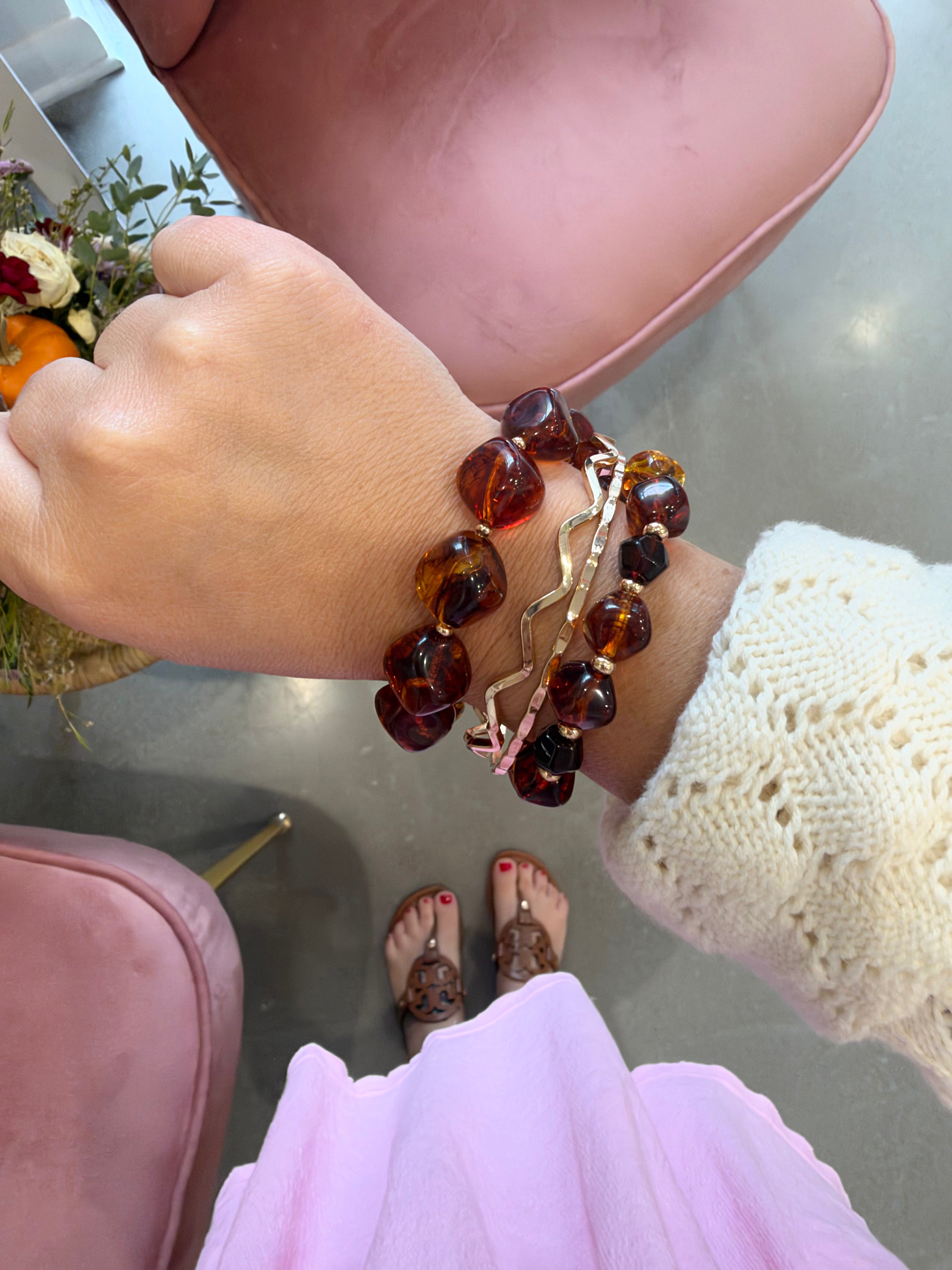 A close-up of a person's wrist wearing multiple bracelets, including amber-colored stone beads and a rose gold chain, with a cream knit sweater sleeve visible. The person is sitting in a pink chair in an indoor setting.