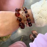 A close-up of a person's wrist wearing three bracelets: two amber-colored beaded bracelets and one gold-toned wavy charm bracelet with a pink accent, shown in a lifestyle setting with a pink chair and gray floor visible.