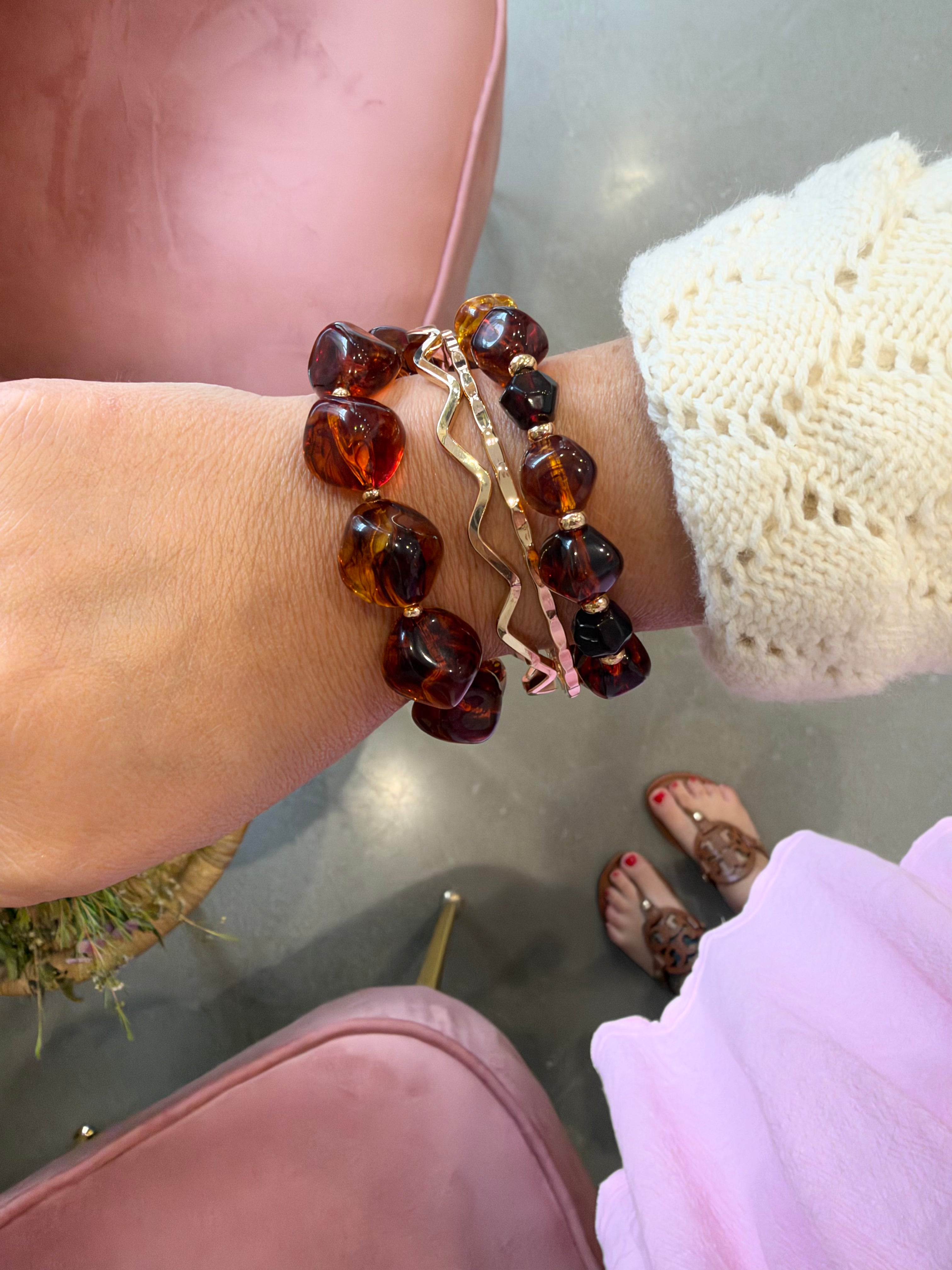 A close-up of a person's wrist wearing three bracelets: two amber-colored beaded bracelets and one gold-toned wavy charm bracelet with a pink accent, shown in a lifestyle setting with a pink chair and gray floor visible.