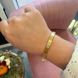 A gold-toned metal cuff bracelet with a small clear gemstone, worn on a person's wrist. The background features a pink velvet chair and a decorative table with flowers.
