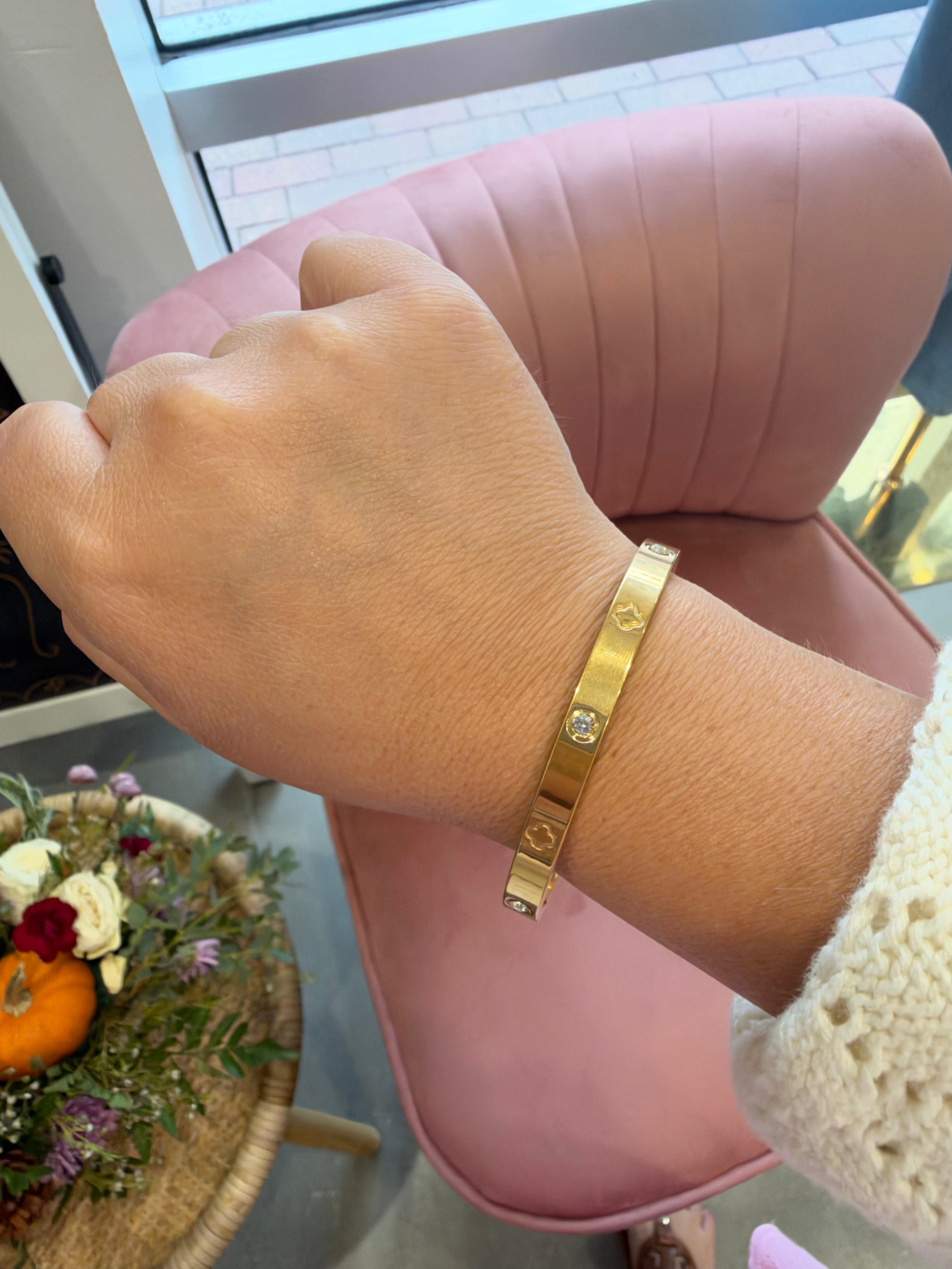 A gold-toned metal cuff bracelet with a small clear gemstone, worn on a person's wrist. The background features a pink velvet chair and a decorative table with flowers.