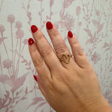 A close-up of a hand with red nail polish wearing a gold ring with two interlocking heart shapes, set against a light wallpaper with a muted floral pattern.