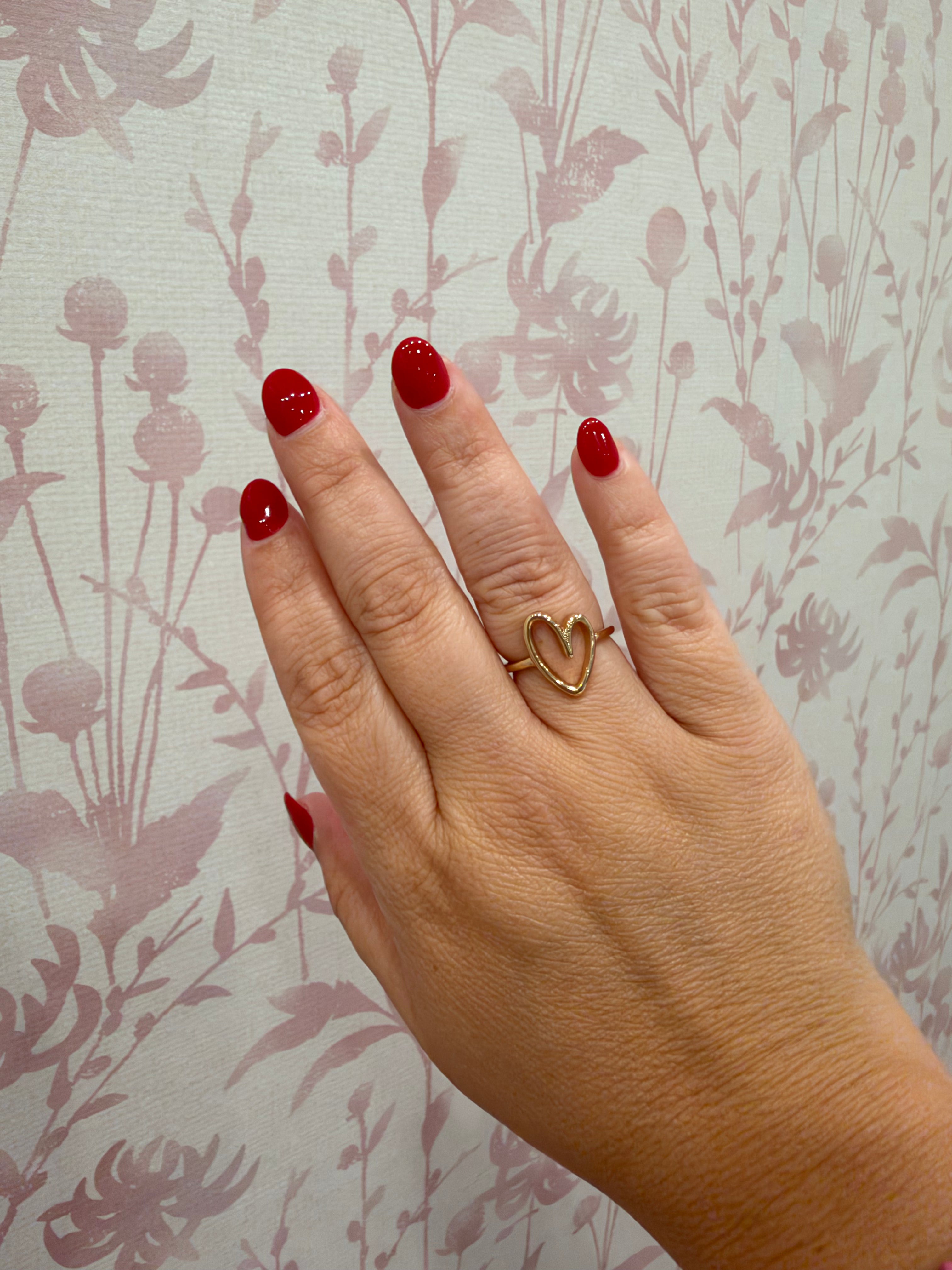 A close-up of a hand with red nail polish wearing a gold ring with two interlocking heart shapes, set against a light wallpaper with a muted floral pattern.