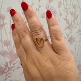 A gold ring with an open heart design worn on a hand with red nail polish, set against a light pink floral patterned background.