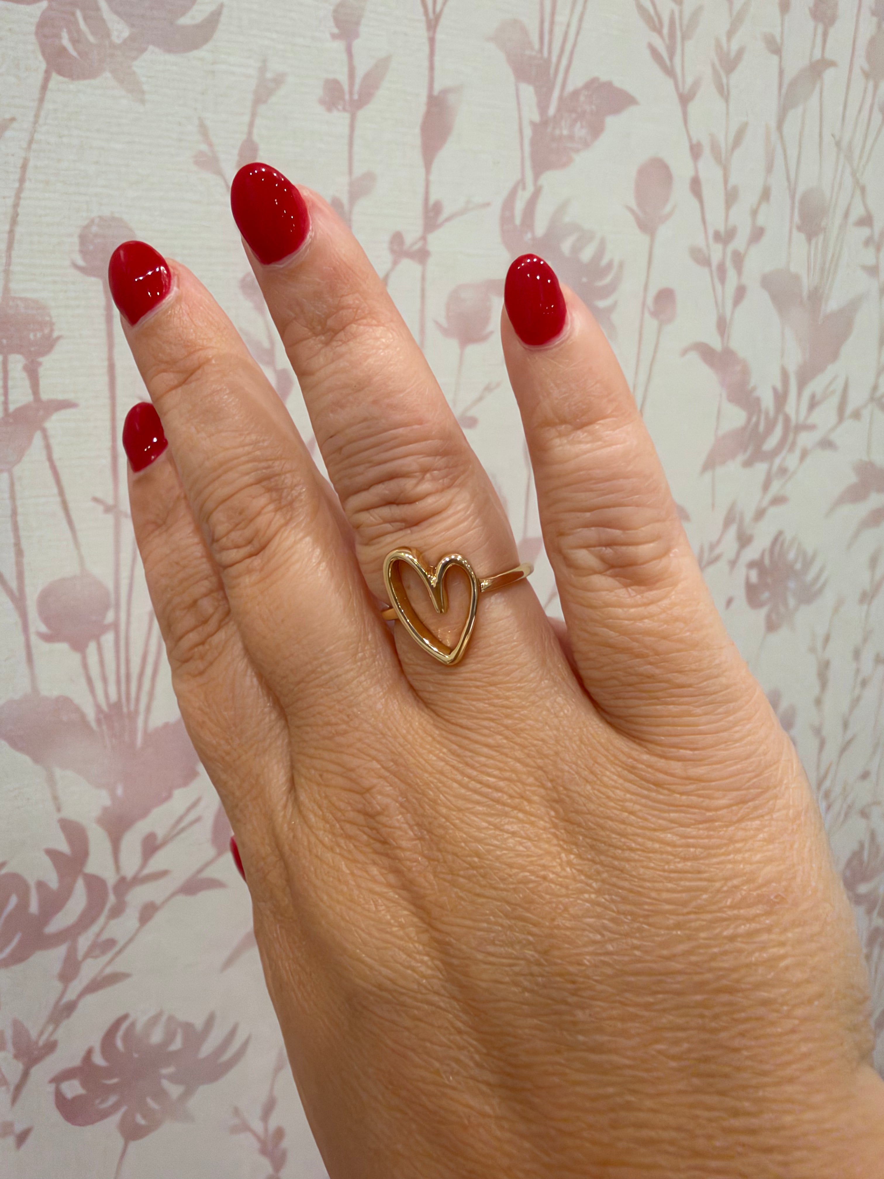 A gold ring with an open heart design worn on a hand with red nail polish, set against a light pink floral patterned background.