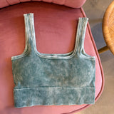 A ribbed knit crop top in a muted sage green color with a faded, washed texture, laid flat on a pink upholstered chair.