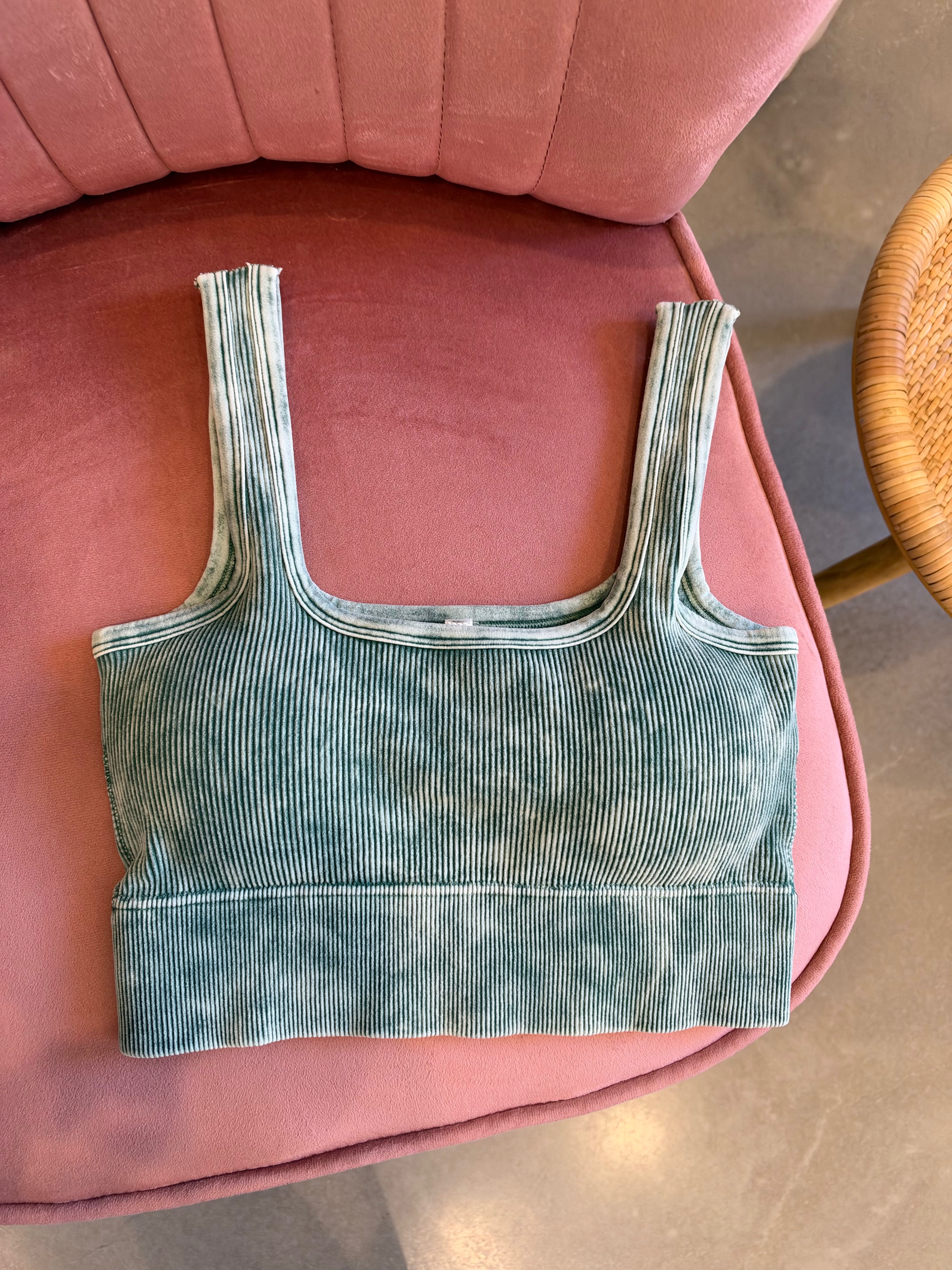 A ribbed knit crop top in a muted sage green color with a faded, washed texture, laid flat on a pink upholstered chair.