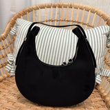 A black crescent-shaped shoulder bag with a curved top handle and silver zipper, displayed on a woven rattan chair with a striped pillow in the background.