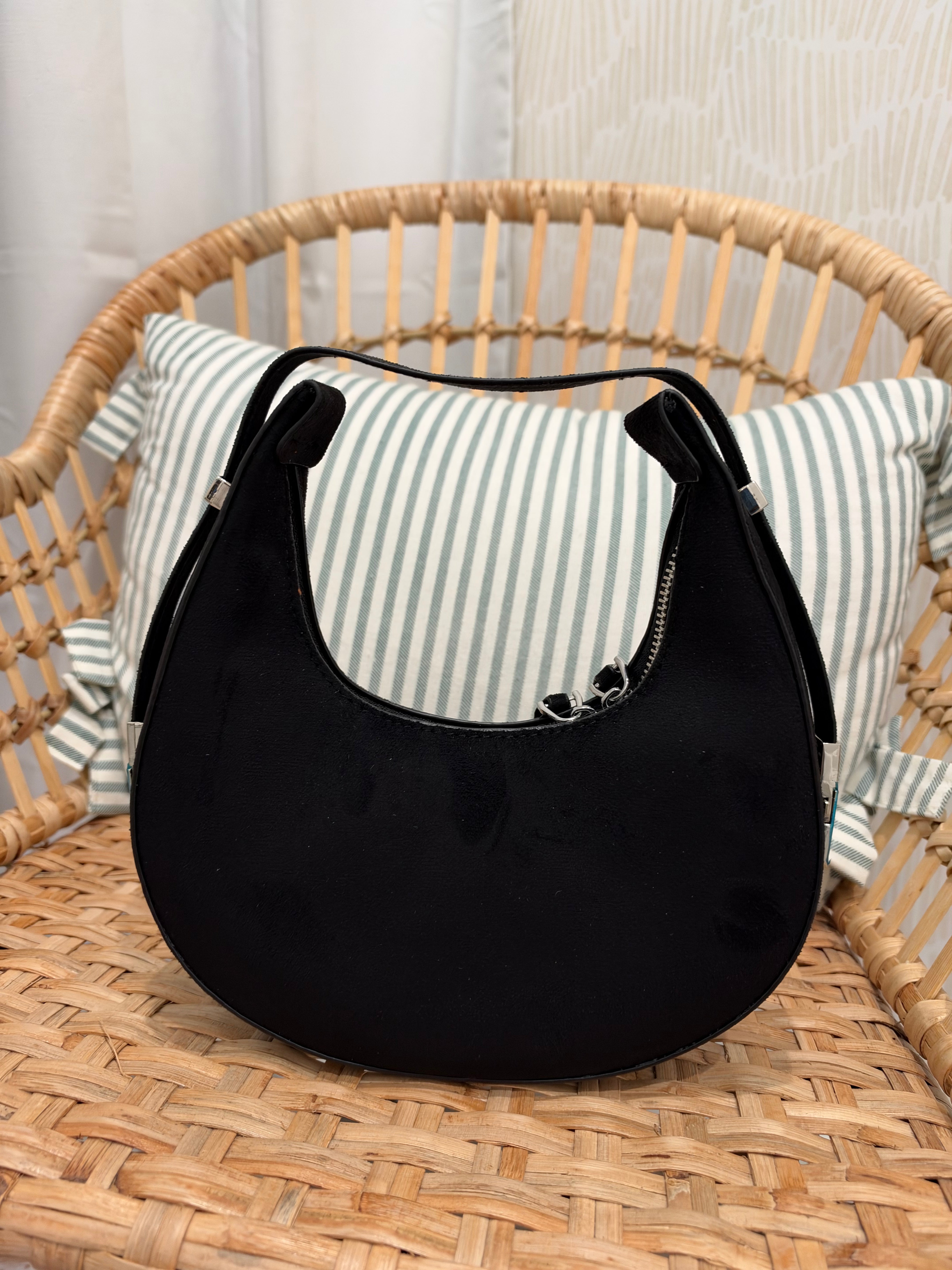 A black crescent-shaped shoulder bag with a curved top handle and silver zipper, displayed on a woven rattan chair with a striped pillow in the background.