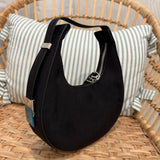 A black crescent-shaped shoulder bag with a top zipper and silver-tone hardware, resting on a woven rattan chair in front of a striped pillow.