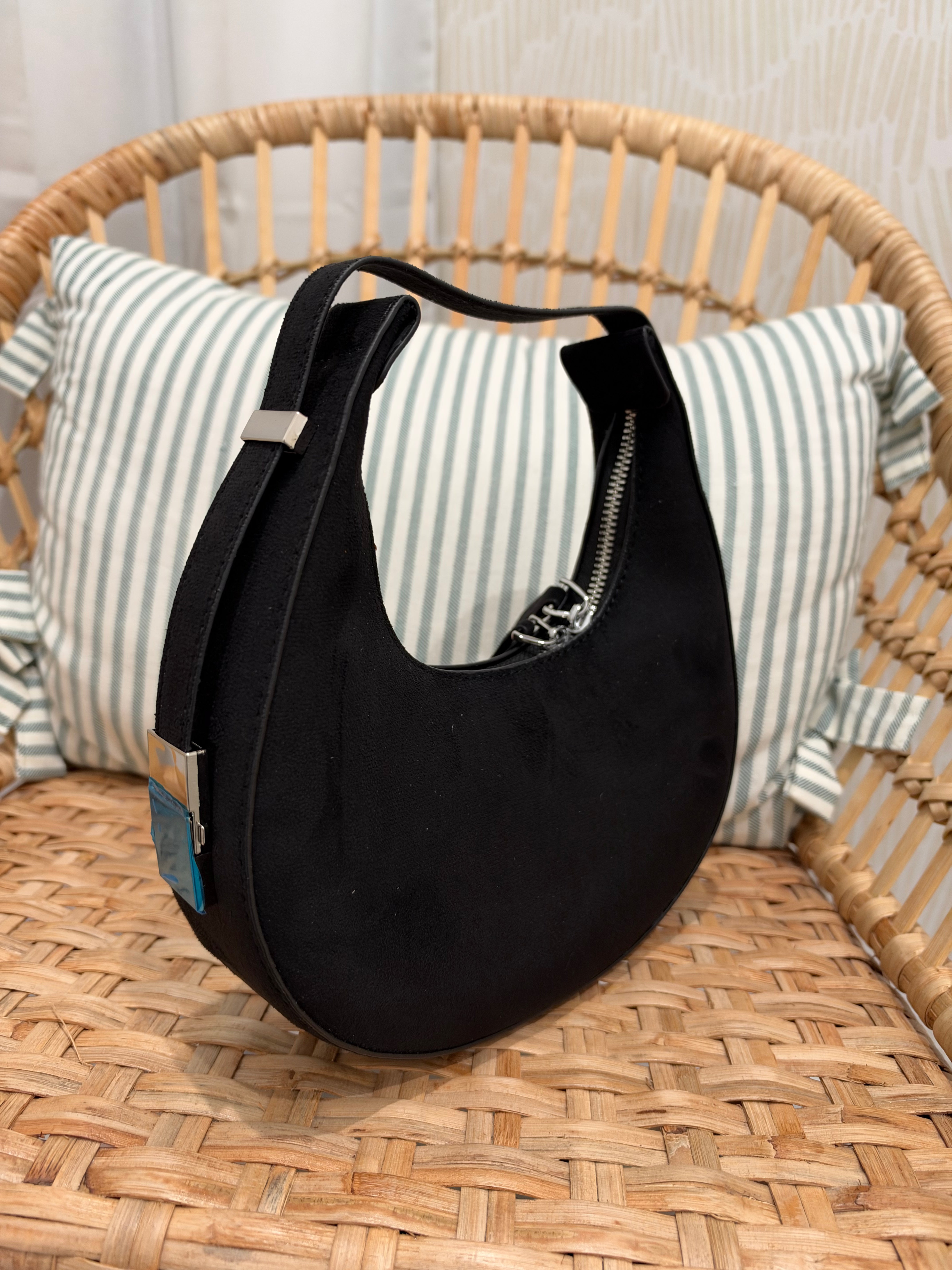 A black crescent-shaped shoulder bag with a top zipper and silver-tone hardware, resting on a woven rattan chair in front of a striped pillow.