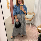 A woman takes a mirror selfie in a fitting room, wearing a blue denim shirt tied at the waist with a black and white gingham maxi skirt, holding a small black handbag.
