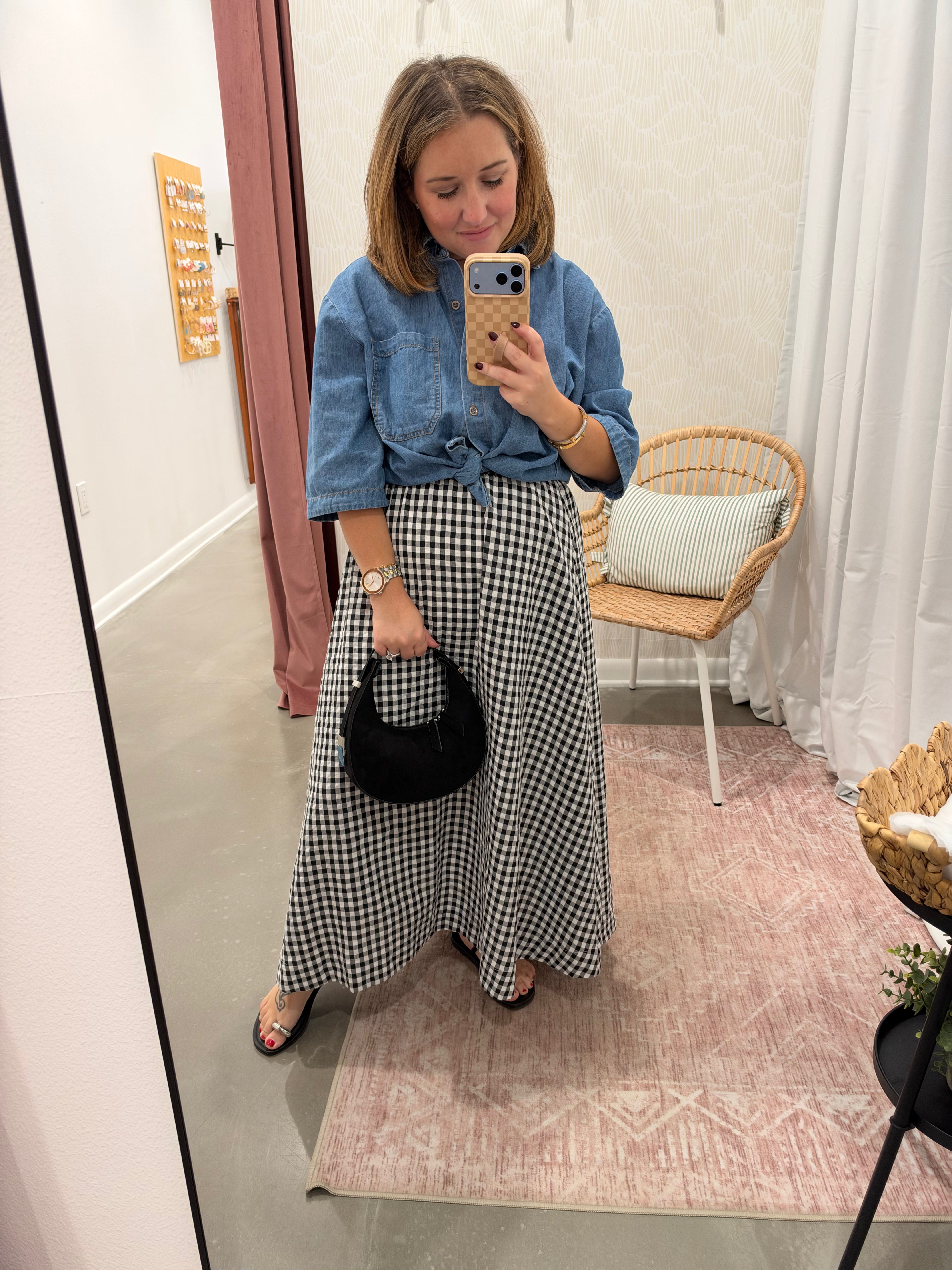 A woman takes a mirror selfie in a fitting room, wearing a blue denim shirt tied at the waist with a black and white gingham maxi skirt, holding a small black handbag.