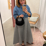 A woman takes a mirror selfie in a fitting room wearing a blue denim shirt and a black and white gingham maxi skirt, holding a black crescent handbag.