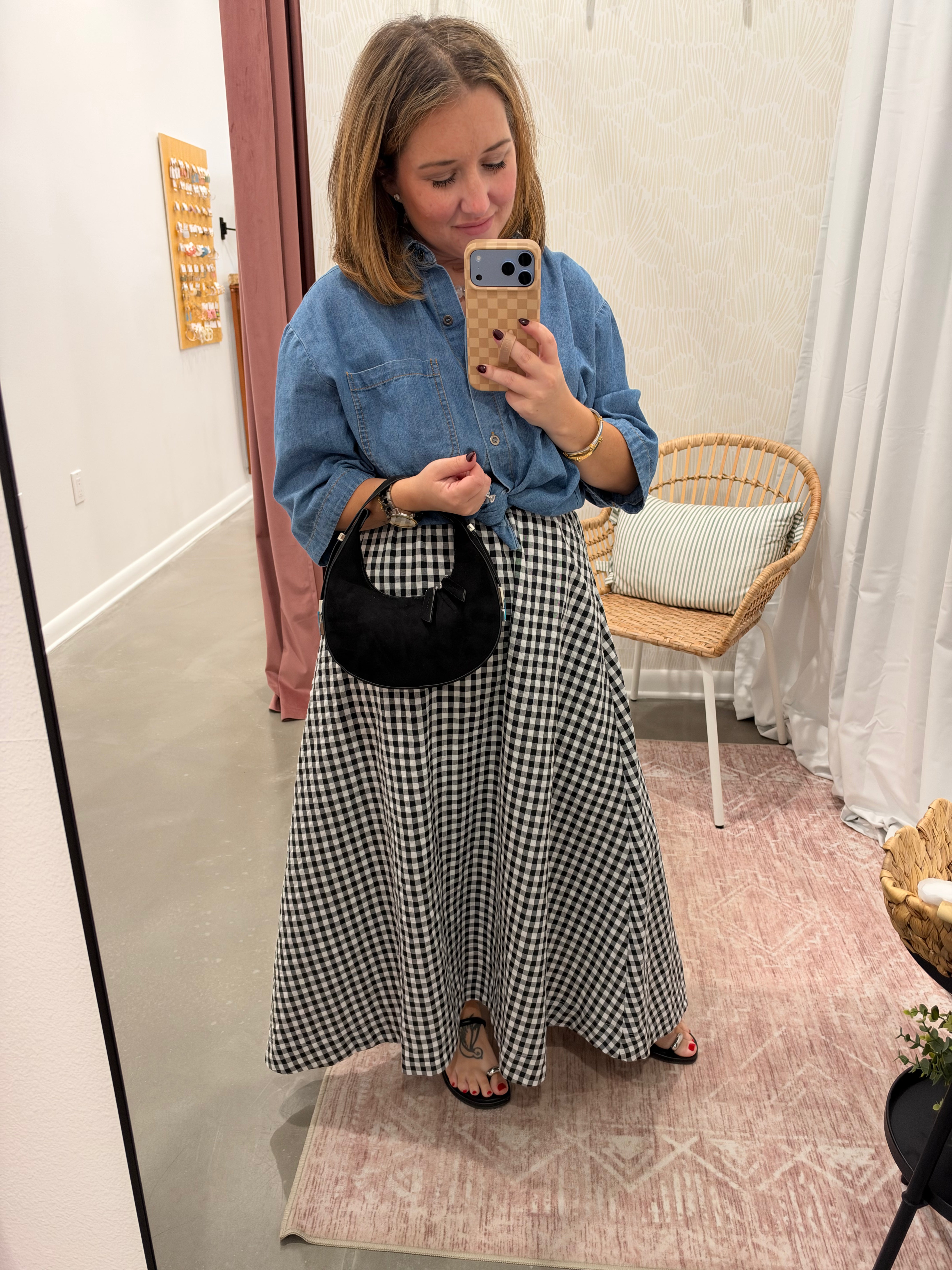 A woman takes a mirror selfie in a fitting room wearing a blue denim shirt and a black and white gingham maxi skirt, holding a black crescent handbag.