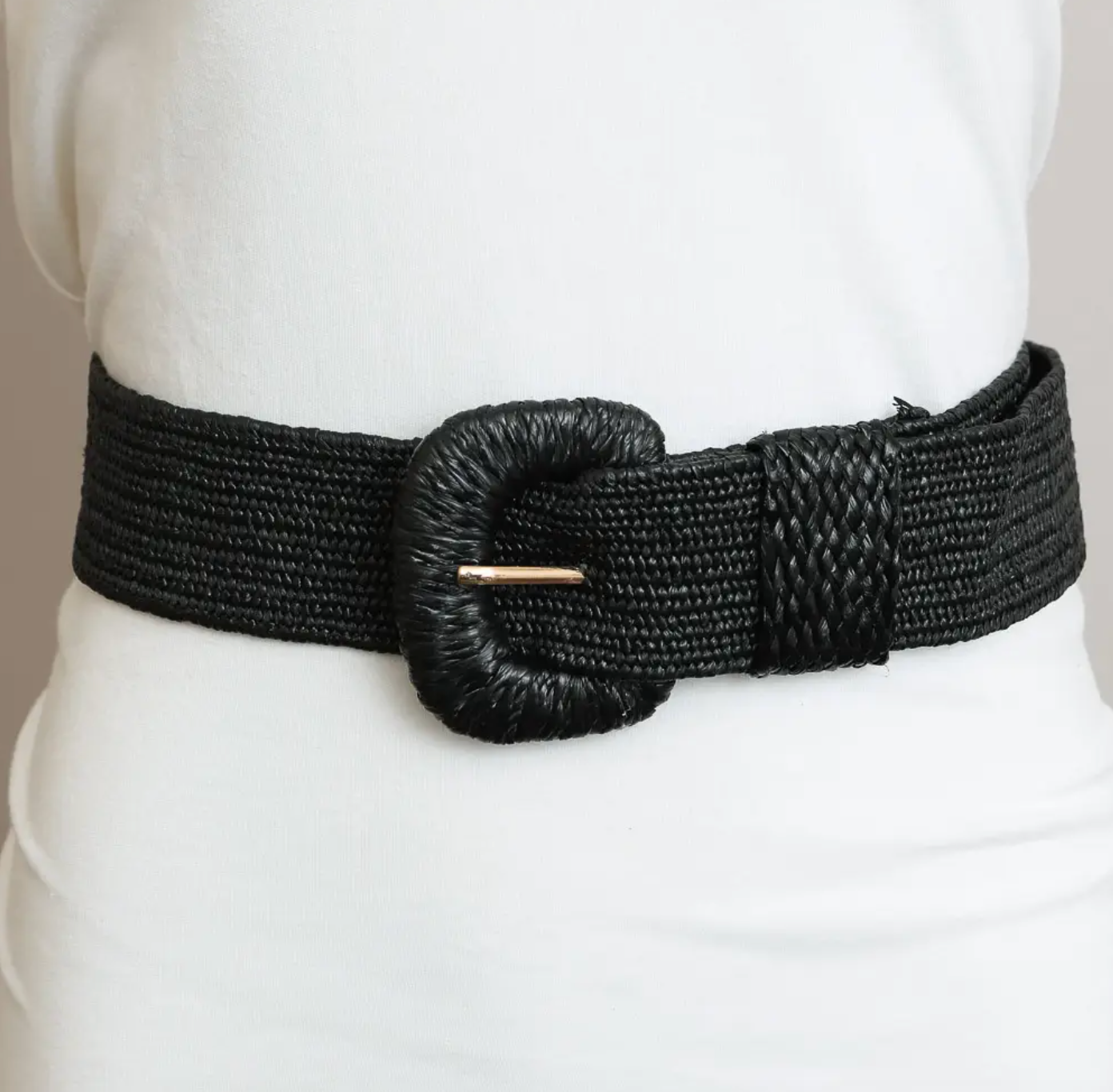 A wide black woven belt with a large textured oval buckle and a small gold-toned bar detail, shown worn over a white top.