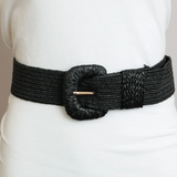 A wide black woven belt with a large textured oval buckle and a small gold-toned bar detail, shown worn over a white top.