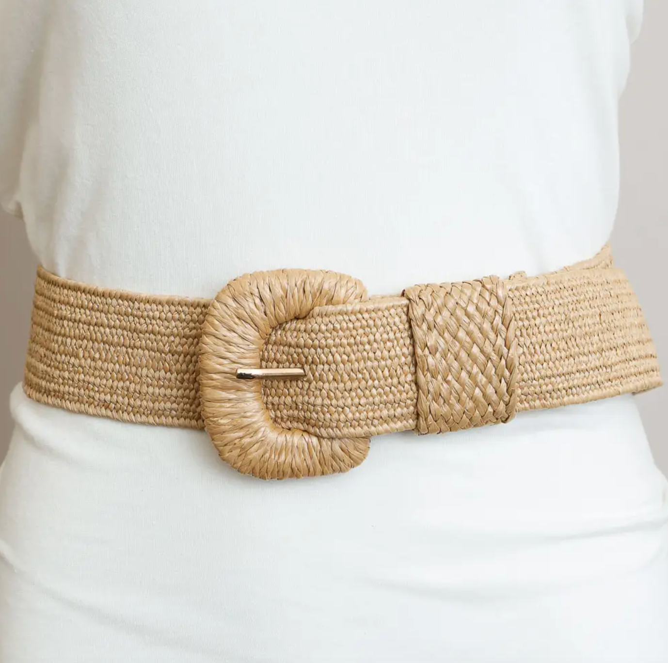A wide woven natural-fiber belt with a large oval buckle, worn over a white top. The belt has a textured braided section and a smooth woven strap, with a small gold-tone buckle.