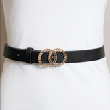 A black faux-leather belt with a double interlocking oval buckle in a gold-tone finish, displayed on a white mannequin torso.